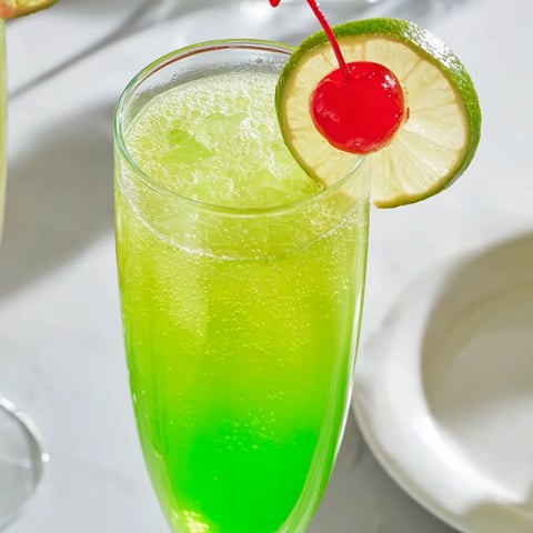 Bright green Quick Grinch Mimosa Mocktail, bubbly and chilled, garnished with a red cherry and lime.